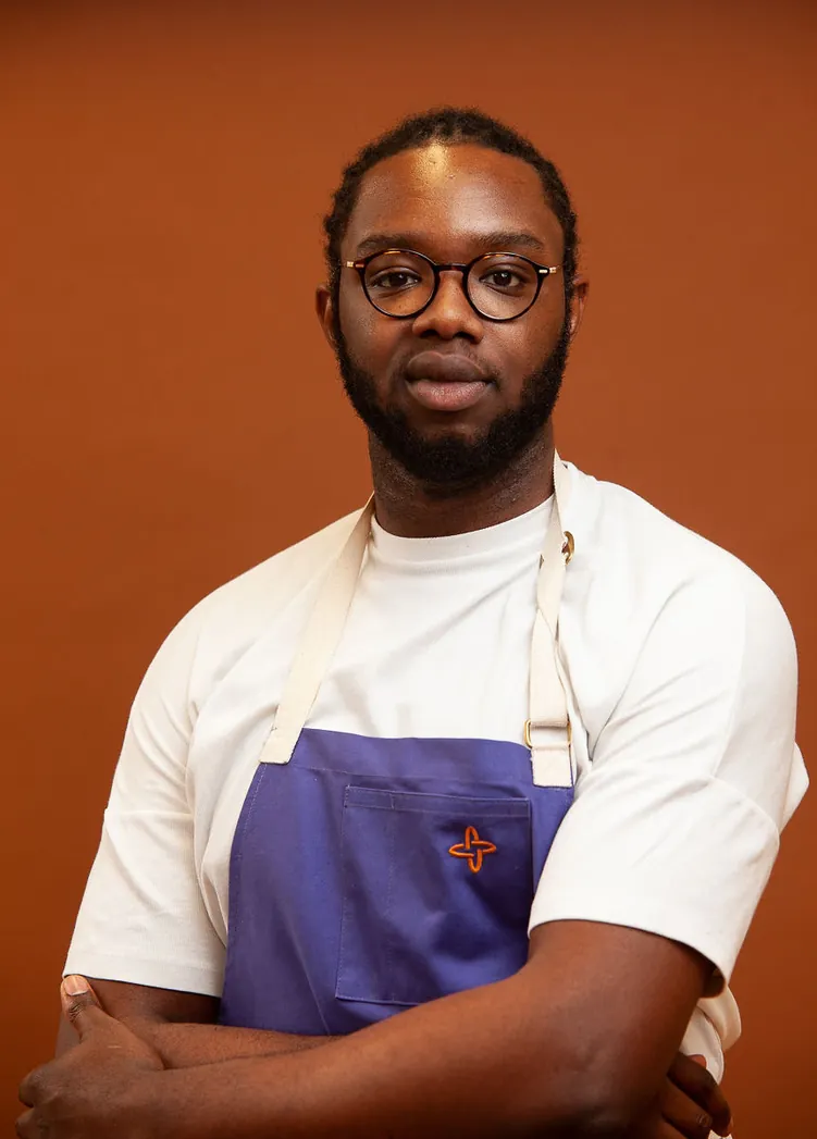 Tobi, the cook behind Kiki, in a purple apron against a burnt orange wall